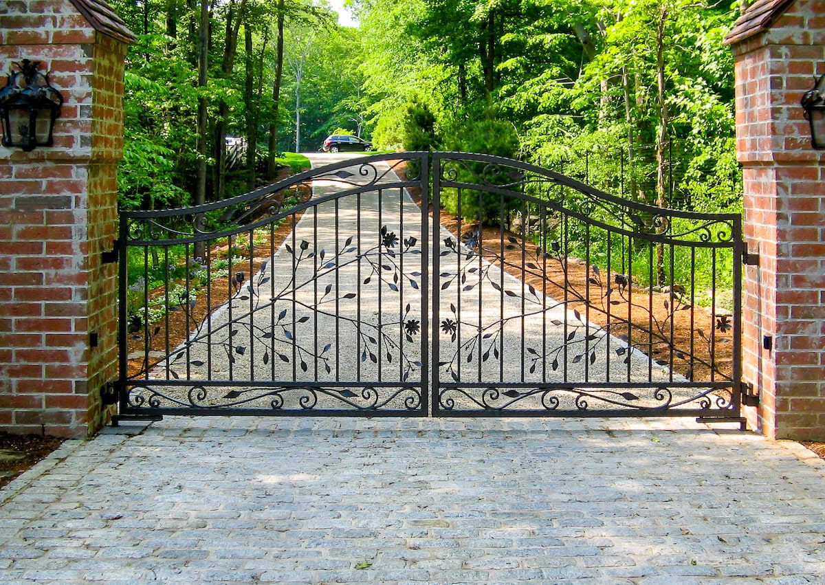 A wrought iron gate with ornate leaf and vine decorations guards the entrance to a long driveway.