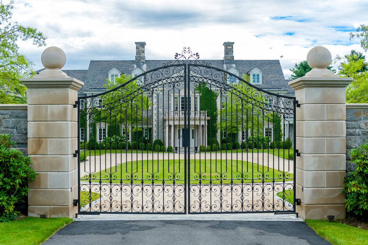 A tall, finely-textured wrought iron gate stands at the entrance to a home with a facade of gray stone and ivy climbing the walls.