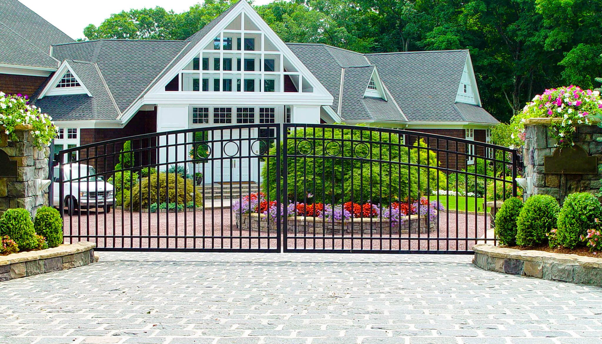  A wide iron gate sits between two stone posts. There is a circular flagstone drive behind the gate. The landscaping is semi-formal, combining shaped shrubs and overflowing flowers.