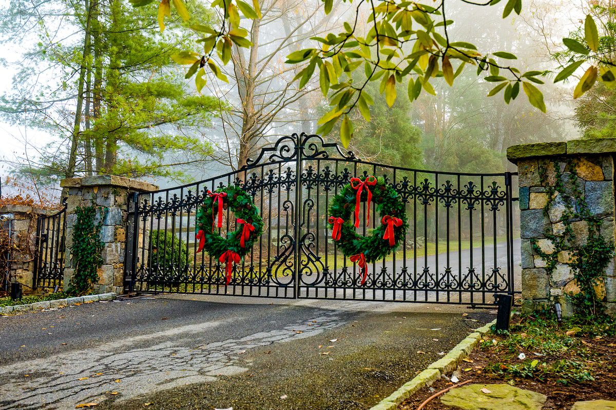 A wrought iron gate with two large Christmas wreaths stands between two stone columns on a foggy day.