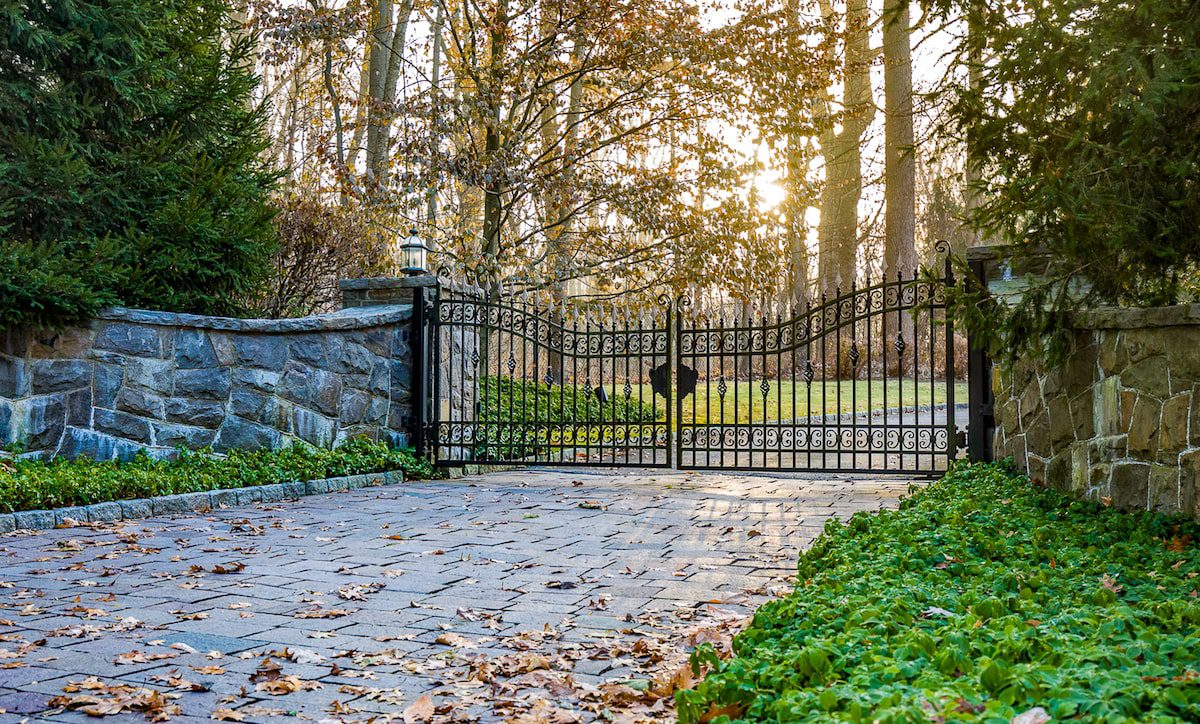 An ornate iron gate sits between two stone posts. The drive is stone, and is bordered by a low groundcover plant.