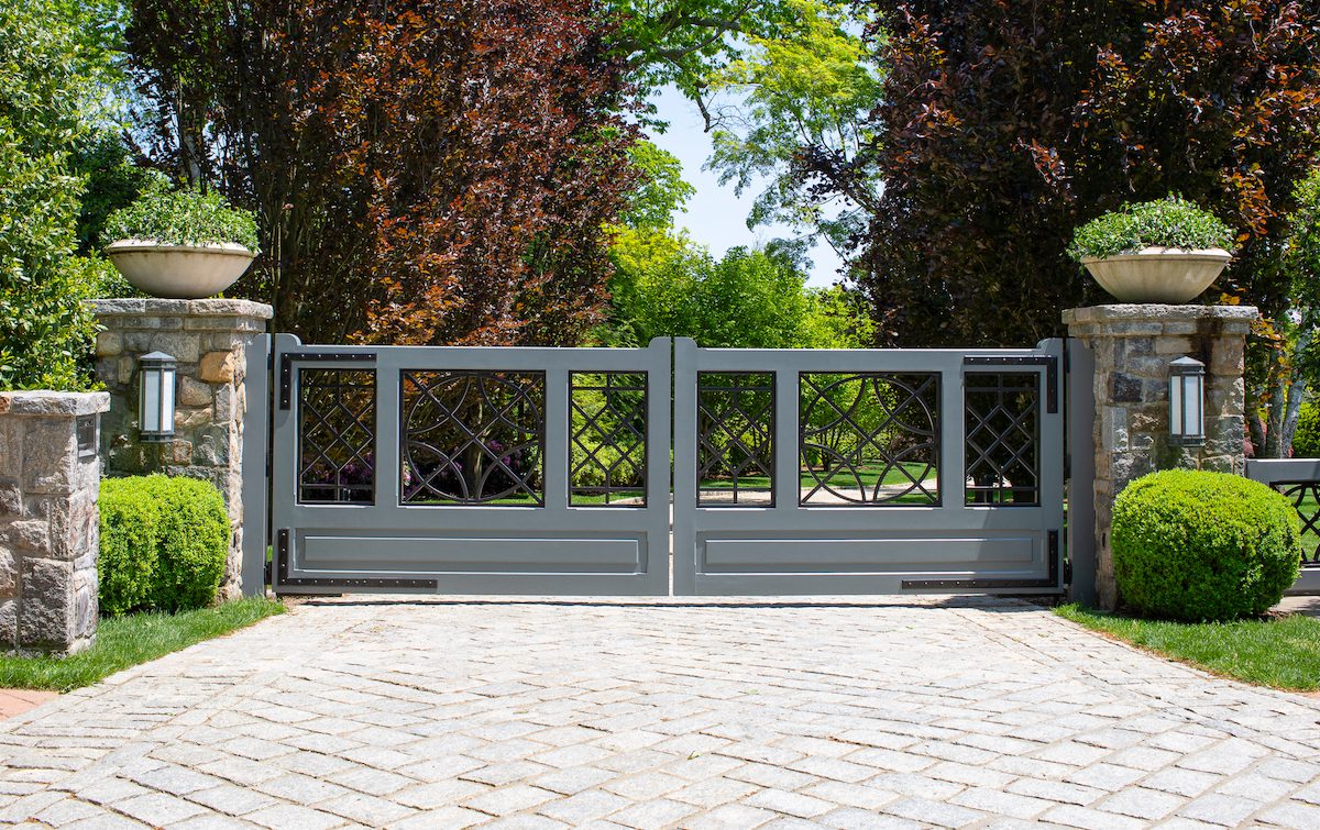 A stylish wood and metal gate sits between two stone posts. There are two ornamental stone bowls filled with greenery sitting on top of the gateposts.