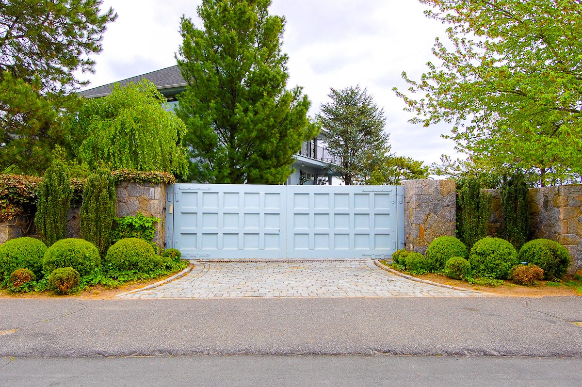 A pale blue composite gate sits between two stone posts. There are shrubs in ascending height order planted along the short flagstone drive between the main road and the gate.