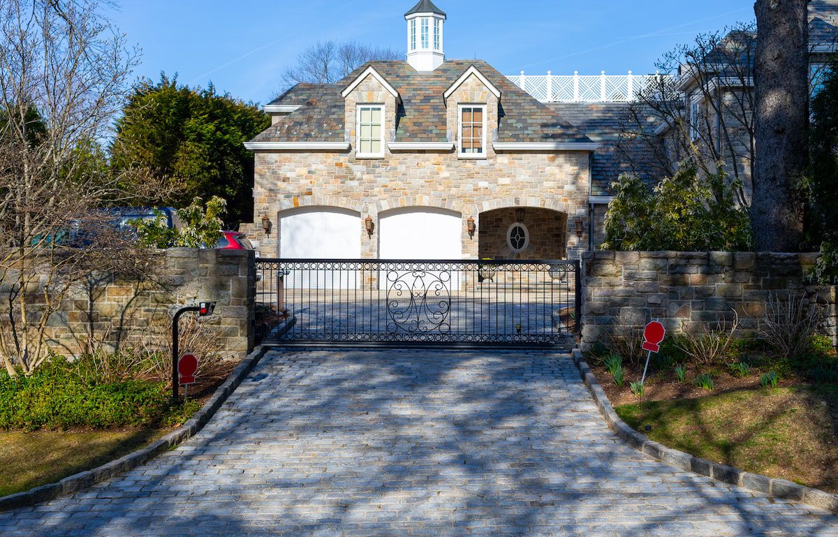 A sliding wrought iron gate guards a stone driveway leading to a two car garage on the side of a home.