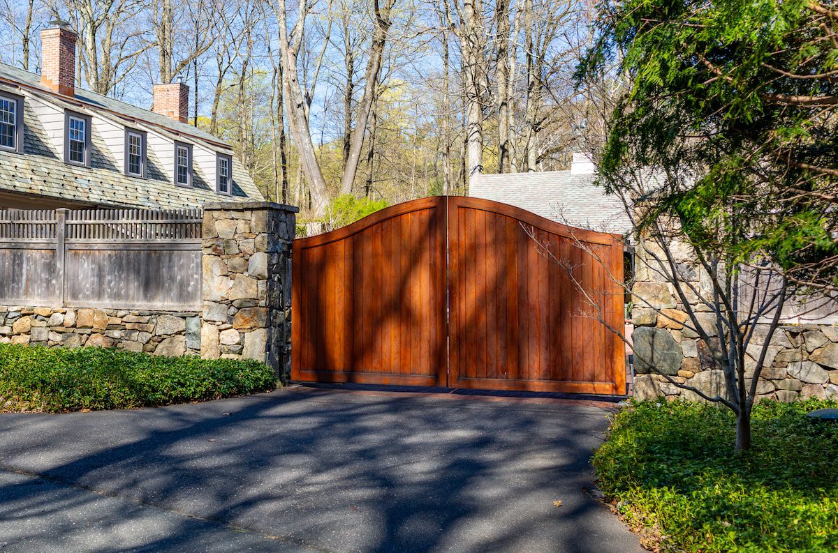 A dark-stained mahogany automatic driveway gate set between stone columns guards the entrance to a home in the background.