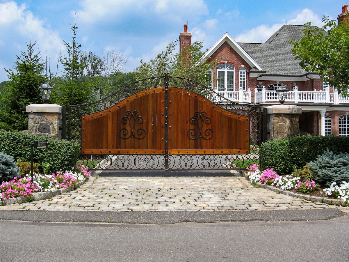 A metal gate contains solid wood panels surrounded with ornamental ironwork. There is an imposing brick house in the background.