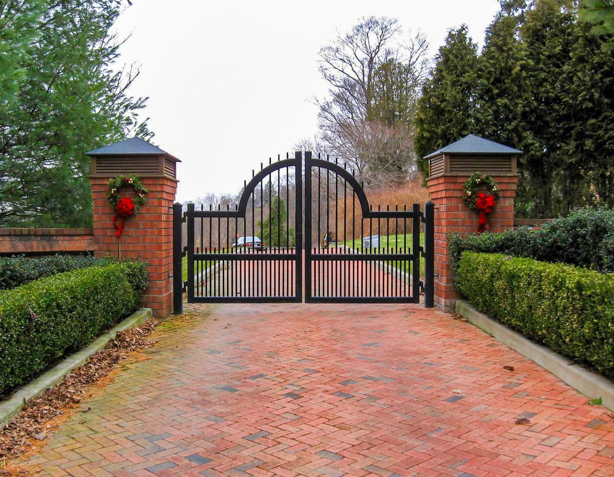 Evergreen shrubs frame a brick drive that leads to an arched iron gate between two brick pillars.