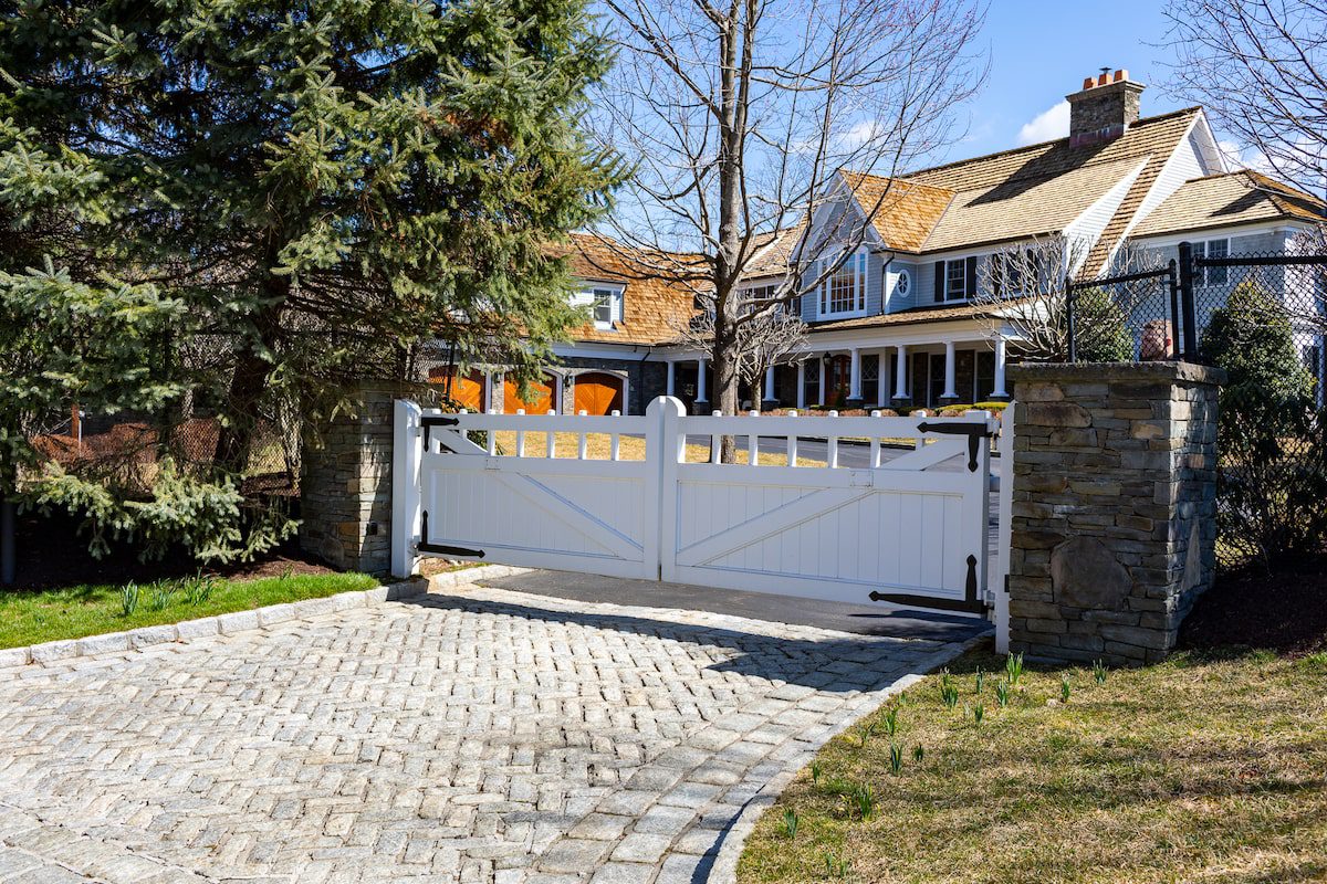 A low wooden automatic driveway gate guards the cobblestone entrance to a large home with a long row of porch columns and three dark-stained wood garage doors.