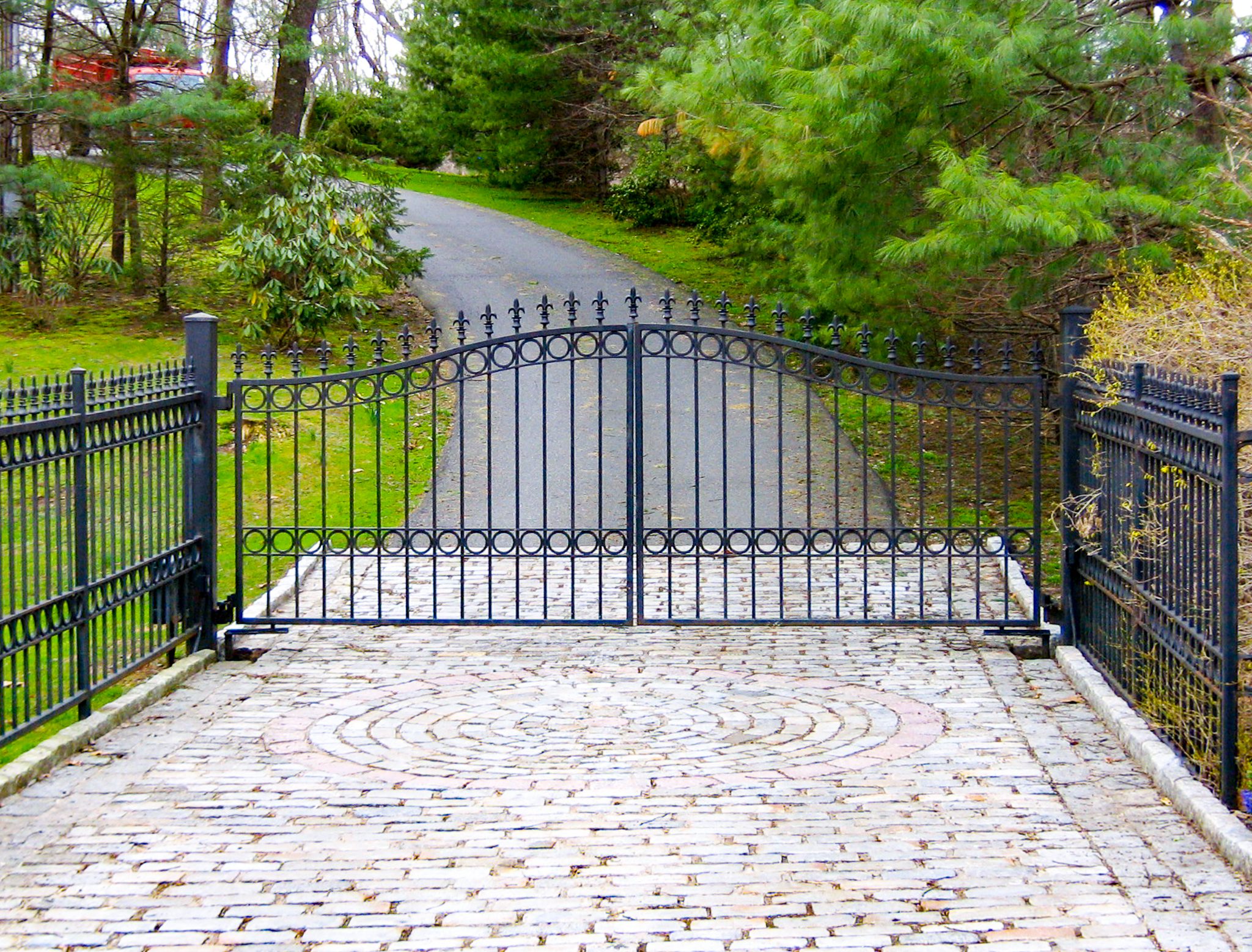 A wrought iron gate with circle designs and fleur-de-lis finials blocks the stone-paved entrance to a property.