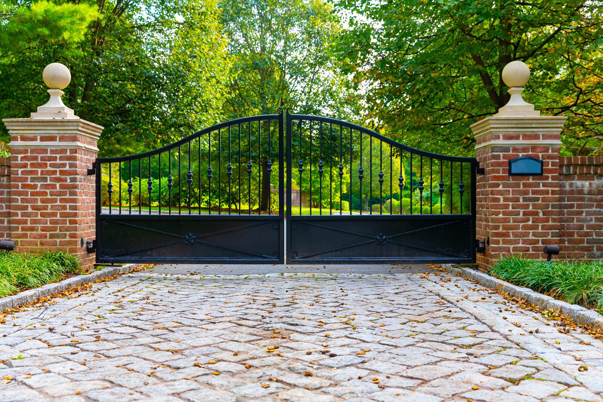 An arched wrought iron swing gate with solid base panels and pickets on the upper portion stands between two wide red brick columns.