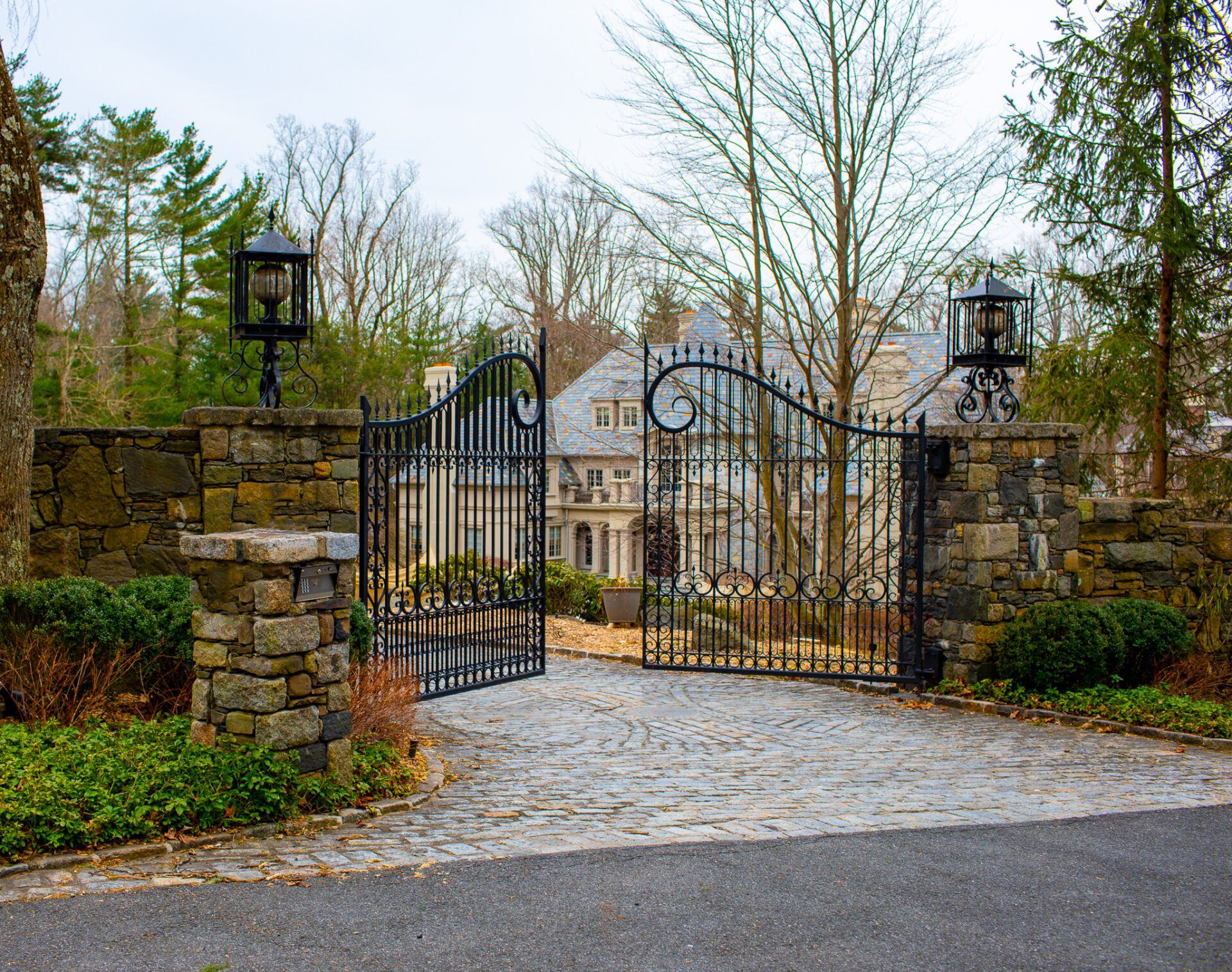 An ornately decorated wrought iron gate with a dramatically arched and curled top rail swings open to reveal a mansion in the background.