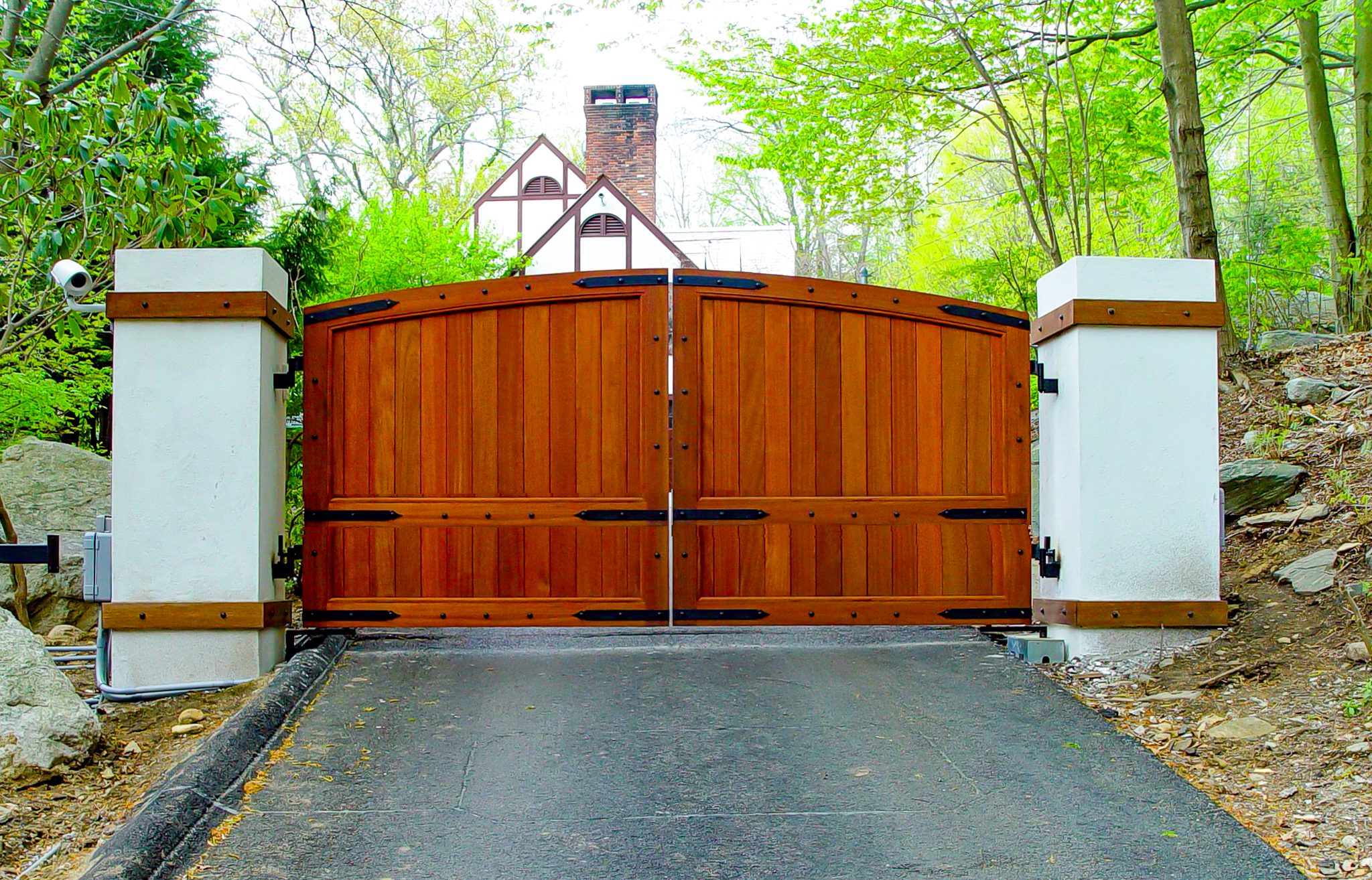 A solid mahogany automatic driveway gate with metal trim on the panels is positioned between two large white stone columns and flanked by a security camera.